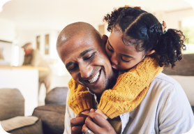 A man sitting on the couch while a little girl wraps around him kissing his cheek while he smiles. 